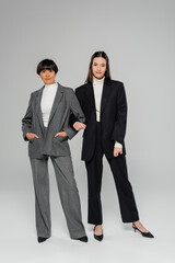 full length of positive asian mother and daughter in elegant pantsuits looking at camera on grey background