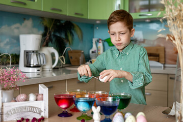 A beautiful red -haired boy paints eggs in the kitchen for Easter. Emotions of surprise. Multi -colored eggs, the child paints at home. Close -up portrait