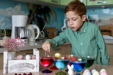 A beautiful red -haired boy paints eggs in the kitchen for Easter. Emotions of surprise. Multi -colored eggs, the child paints at home