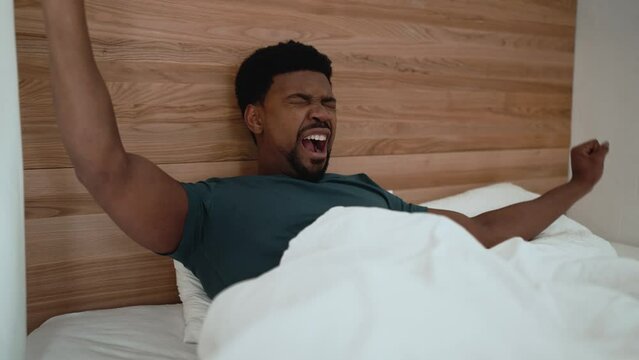 Handsome African brunet man yawning and stretching while lying on the bed at home
