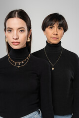 trendy asian mother and daughter in black turtlenecks and necklaces looking at camera isolated on grey