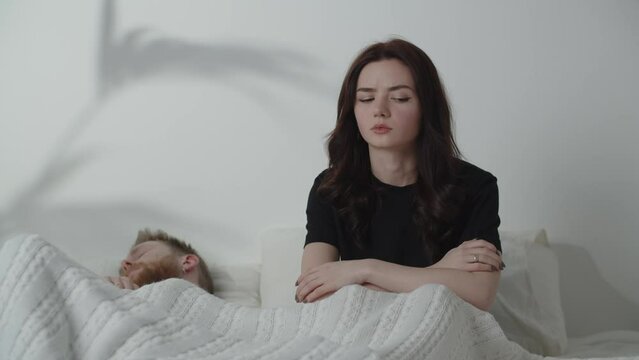 Disappointed girl looks at her sleeping boyfriend. Sexual problems idea. Relationship difficulties. Young woman sitting on bed and thinking about divorce