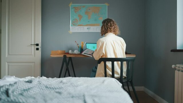 Back view of curly haired concentrated woman studying with books and a laptop at the table at home