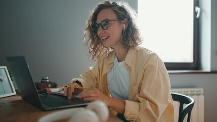 Side view of curly haired cheerful woman talking by video call on laptop at the table at home - Powered by Adobe