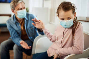 Distraught little girl and her father waiting for medical exam at doctor's office during coronavirus pandemic.