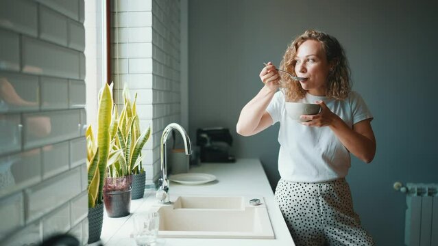 Positive curly haired woman eating cereal with milk and looking at window in the kitchen at home
