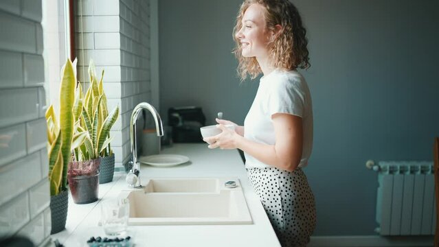 Cute curly haired woman eating cereal with milk and looking at view in the window at home