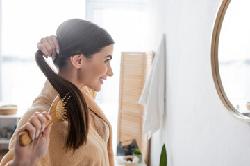side view of happy young woman brushing shiny hair and looking at mirror in bathroom.