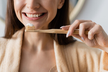 partial view of smiling woman in bathrobe holding toothbrush with toothpaste in bathroom.