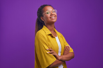Young satisfied ethnic African American woman in casual oversize clothes arms crossed looks at camera and raises chin proud of success in career or own business stands in purple studio