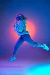 Fototapeta na wymiar Active lifestyle, plans. Young woman in casual clothes, hoodie and jeans running over gradient studio background in blue neon light. Concept of emotions, facial expression, inspiration, sales, ad