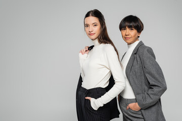 positive asian mother and daughter in formal wear posing with hands in pockets while looking at camera isolated on grey