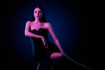 Fototapeta na wymiar Beautiful young caucasian brunnete girl sitting on chair in sexy black dress in colored blue purple light in studio on a dark background