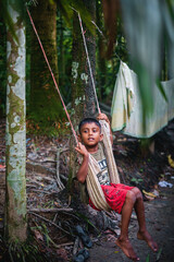 Little Bangladeshi kid playing with a swing made with clothes and rope using trees as support in a village garden  - obrazy, fototapety, plakaty