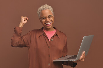Young overjoyed attractive African American woman freelancer holding open laptop and waving after receiving new lucrative order or learning how to do work more efficiently stands on brown background