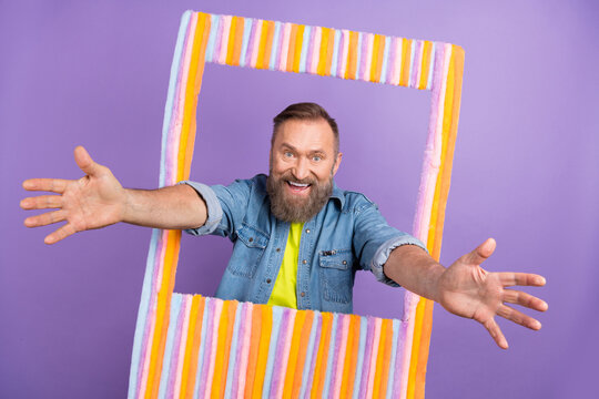 Portrait of positive aged man raise opened arms through paper album card photozone invite you isolated on purple color background
