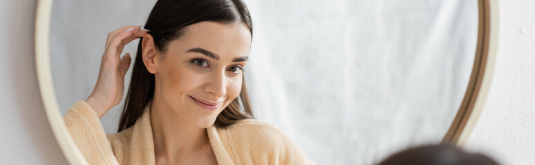 young brunette woman adjusting hair and smiling while looking at mirror in bathroom, banner.
