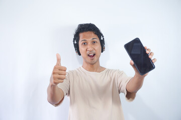 Half length shot of optimistic man holds smartphone with mock up screen, listens favourite soundtrack in headphones, wears casual cream t shirt, stands indoor. People and technology concept.