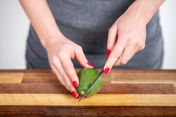 Female hands with red nails cutting a green avocado on a wooden board. Fresh raw vegan salad preparation. - 574281863
