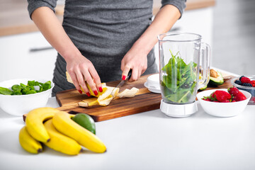 Woman with red nails chopping banana when preparing healthy smoothie. Raw drink made of banana, berries, avocado and spinach.