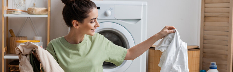 Smiling woman holding white and colorful clothes in laundry room, banner. - obrazy, fototapety, plakaty