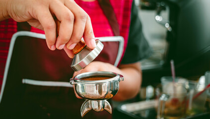 Hand of a barista holding a portafilter and a coffee tamper making an espresso coffee. Barista presses ground coffee using a tamper in a coffee shop