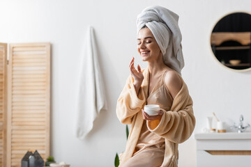 joyful young woman with towel on head holding container with cosmetic cream.