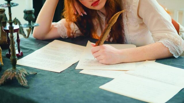 Girl fantasy princess sits at desk writes letter on sheet paper. woman writer holds pen feather quill in hands close-up works signs documents. Beauty face cropped, red lirs long hair. Vintage old book