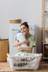 Positive woman holding washing liquid and looking at camera in laundry room.