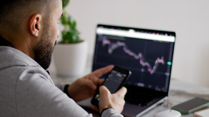Happy bearded young man looks at the screen of his smartphone, sitting in front of laptop screen with graph of securities report. Businessman in casual clothes is working in the office on project.