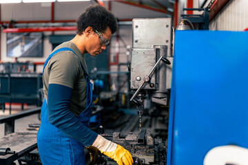 A worker dressed in overalls and protective gloves, wearing safety glasses uses a machine to drill holes in a metal rod