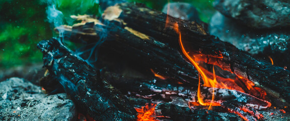 Vivid smoldered firewoods burned in fire close-up. Atmospheric warm background with orange flame of campfire and blue smoke. Wonderful full frame image of bonfire. Burning logs in beautiful fire.