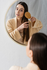 reflection of brunette young woman brushing shiny hair and looking at mirror in bathroom.