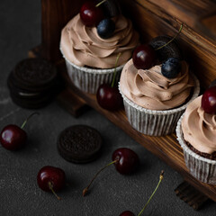Cupcakes with cherries and chocolate cream. Three cupcakes on wooden shelf, decorated with berries, cherries and chocolate biscuits. Festive baking. Dark background. Soft focus. Top view.