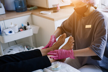 Nail artist in beauty salon making pedicure for client feet. Professional in mask on face polish nails by tool. Concept of nails, feet health and podiatrist profession                             - obrazy, fototapety, plakaty