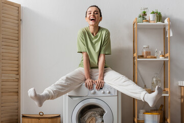 Excited woman sitting on washing machine with clothes at home.
