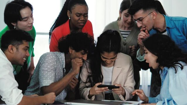 Multiethnic diverse friends playing a game online on smartphone mobile phone, friends support the gamer. Video game addiction. Young international people students having a break, playing on phone 