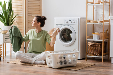 Side view of cheerful brunette woman holding clothes near basket in laundry room.