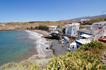 View of the beach and the houses of a small fishing village. White houses on the right and old wooden boats on the shore of the rocky beach. Turquoise ocean on the right, hills in the background.
