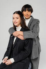 brunette asian woman embracing young and elegant daughter while looking at camera isolated on grey