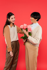 happy asian daughter looking at cheerful mother with flowers in hands isolated on coral