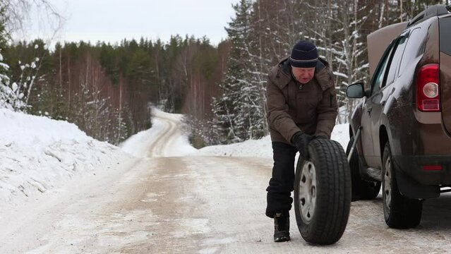  Motorist rolls punctured wheel to trunk of car after installing spare wheel on winter day on forest road.