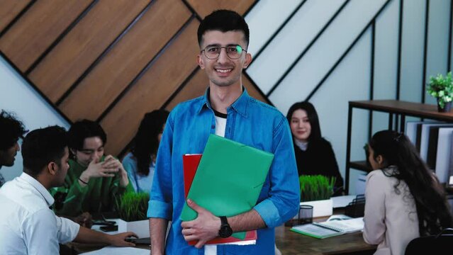 Portrait of Happy smiling middle eastern male student standing in class, holding books and looking at camera. Classmates students are on background. Study at university, education concept. 