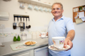 Fototapeta na wymiar Retirement: Tea for Two. Foreground focus on the cup being offered by a smiling senior man in a domestic kitchen. From a series of related images.