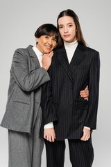 happy asian woman in grey suit hugging young daughter while looking at camera isolated on grey