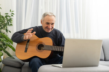 Obraz premium Portrait of senior man in headphones taking online guitar lesson looking at laptop screen. Retired male learning to play guitar watching webinar on computer at home