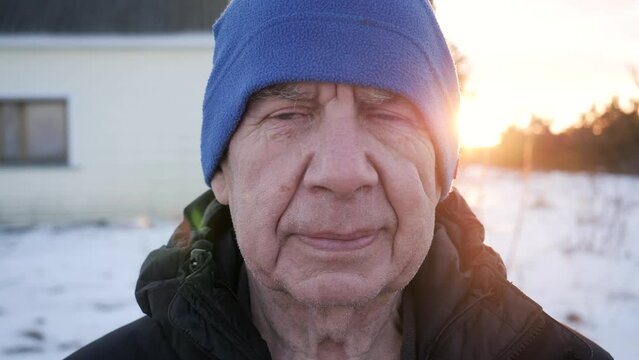 Portrait of alone unhappy elderly old man with deep wrinkles on face. Male looks in camera with calm, sad plaintive look, tears come to eyes. Standing outdoor in winter sunset, walking in fresh air