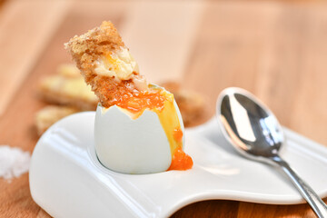 a single boiled egg in an egg cup, broken open showing yolk and with a slice of toast inserted with a spoon on a wooden surface - 574318439