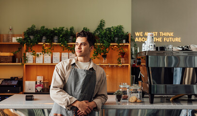 Confident local coffee shop owner leaning counter wearing an apron. Male barista looking away in a cafe.