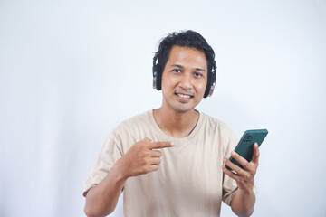 Young smiling fun cool man of Asian ethnicity 20s wearing cream shirt headphones listen to music hold use mobile cell phone isolated on plain white background studio portrait